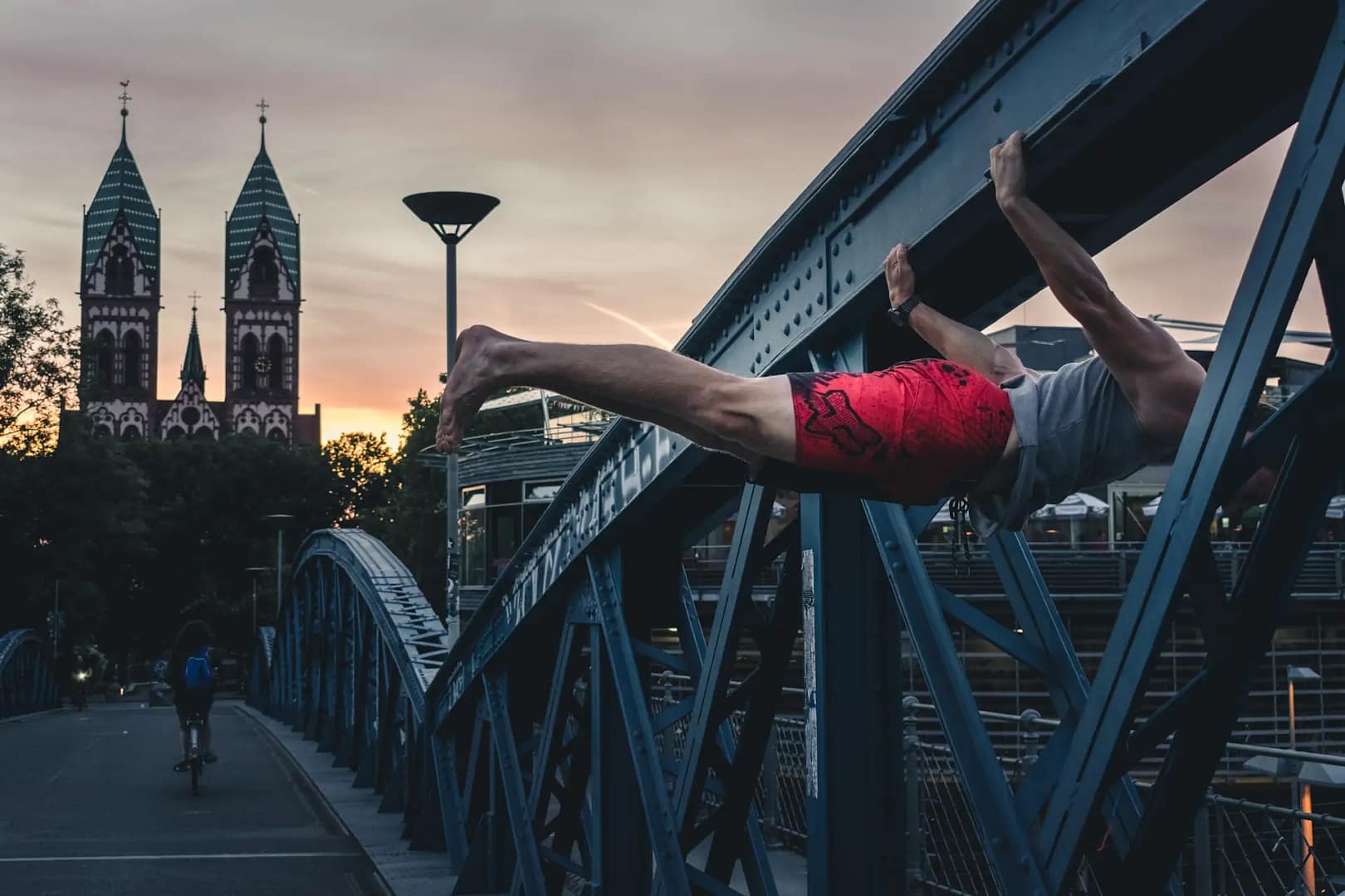 A person performing a back lever on an iron bridge at sunset, with a church in the background and a biker passing by.